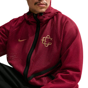 Person wearing a red Nike Chivas 26/27 Third Jacket, a hooded zip-up soccer gear piece with a gold logo on the chest and a matching gold check mark.