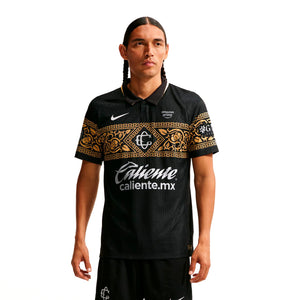A man wears a black Nike Chivas 26/27 Match Third Autenthic Jersey featuring a gold ornate pattern across the chest and sleeves, sponsor logos, and a polo-style collar.