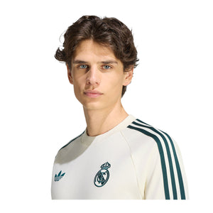 A person wears a cream-colored adidas Real Madrid Originals T-Shirt with dark green three-stripe detailing on the shoulders, a trefoil logo on the right chest, and a soccer club crest on the left chest.