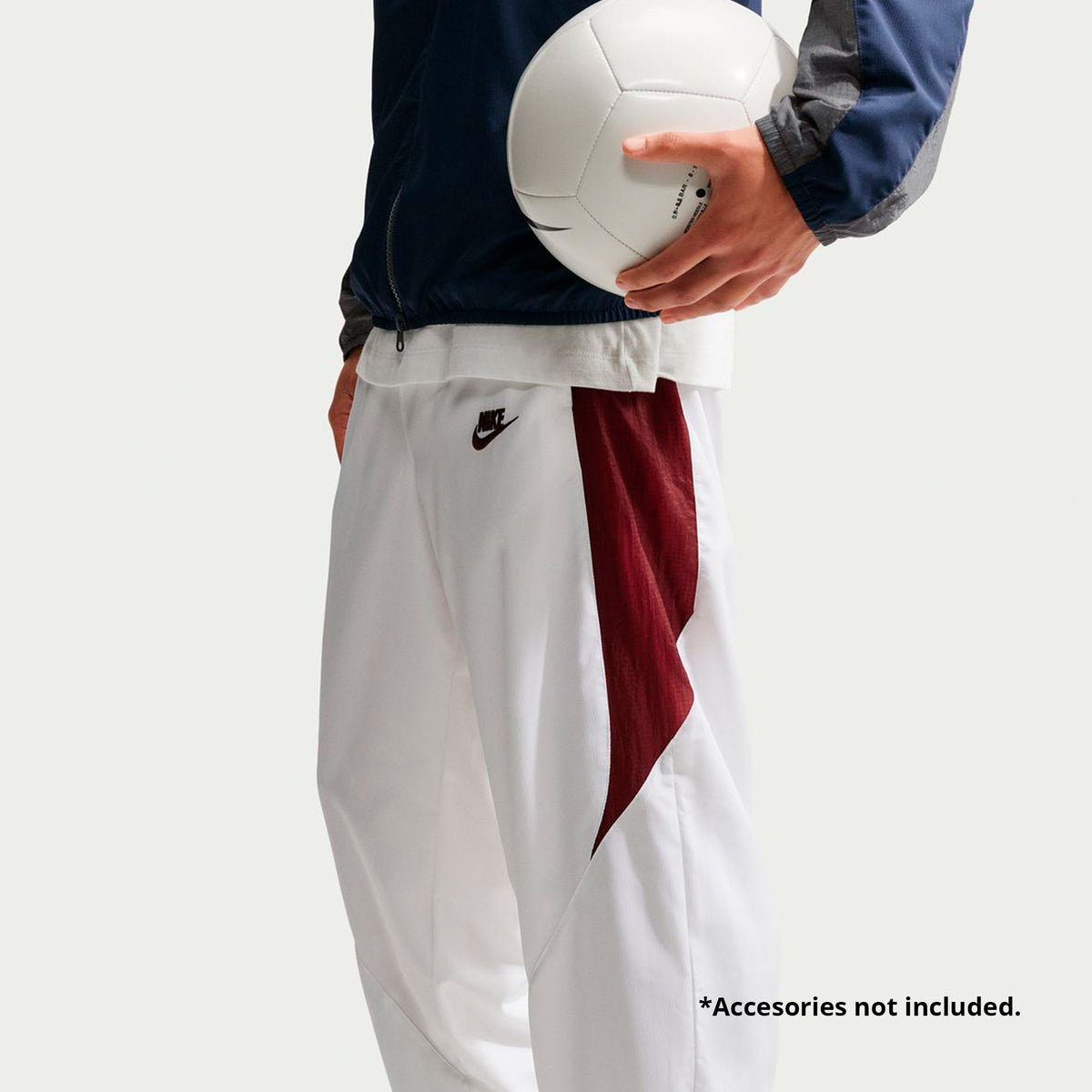A person wearing Nike Premier Repel Pants, which are white with maroon side panels and a dark-colored logo on the upper thigh, holding a white soccer ball.