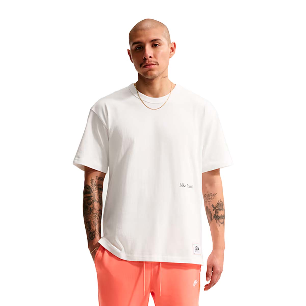 A man wears a white Nike Sportswear T-Shirt with small black text on the chest and a hem patch, paired with bright orange shorts for a complete game-day look.