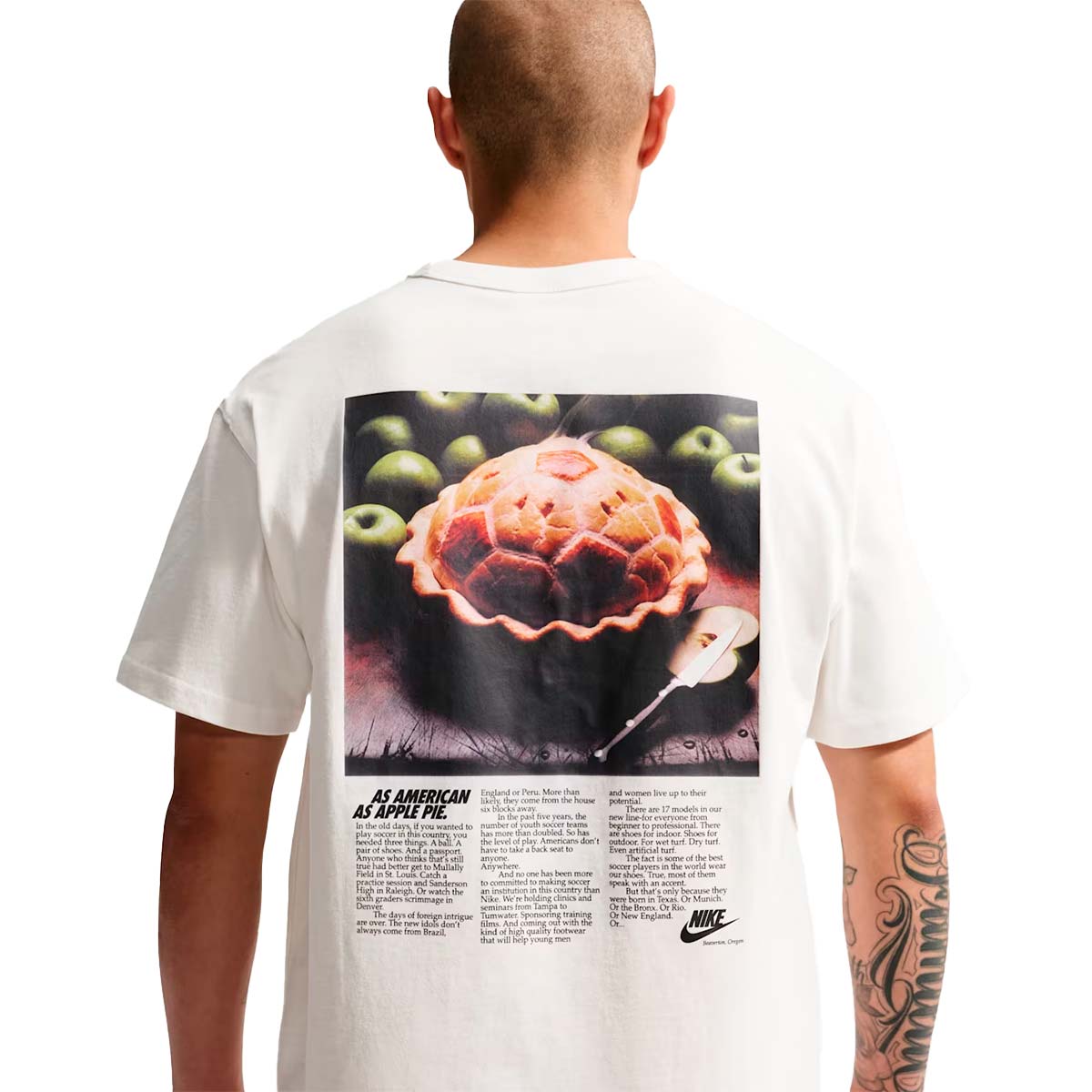 A person seen from behind wears a white Nike Sportswear T-Shirt featuring a large graphic print of a pie surrounded by green apples, with soccer-themed text below the image for a game-day look.