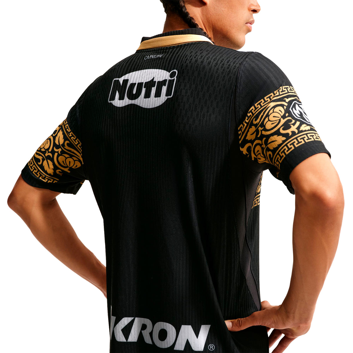 A person wearing a black Nike Chivas 26/27 Match Third Autenthic Jersey featuring gold patterned sleeves and white sponsor logos on the upper back and lower hem.