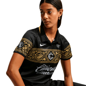 A person wears a black Nike Chivas 26/27 Stadium Third Jersey, featuring a gold filigree band across the chest and sleeves, with white sponsor logos and a team crest.
