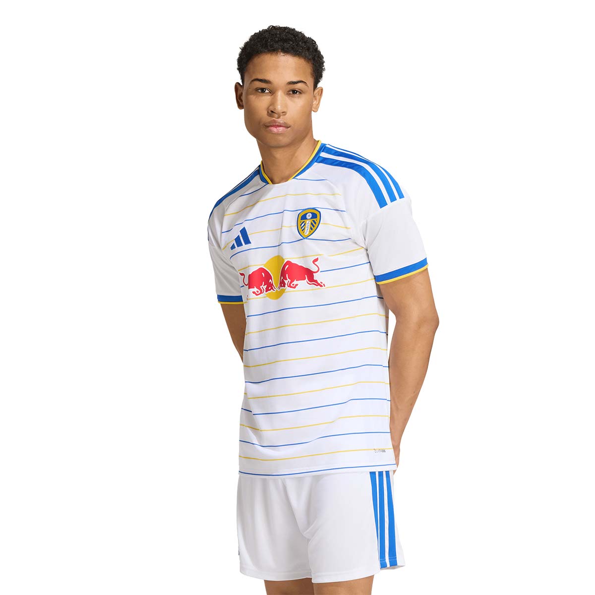 A man wearing an adidas Leeds United 26/27 Home Jersey with thin horizontal blue and yellow stripes, a team crest, and a Red Bull logo on the chest, paired with white shorts.