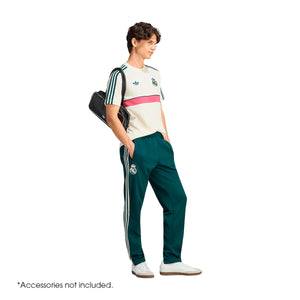 A person wears an adidas Real Madrid Originals T-Shirt in cream with a pink horizontal stripe, paired with dark green sweatpants, both featuring team logos and signature three-stripe detailing.